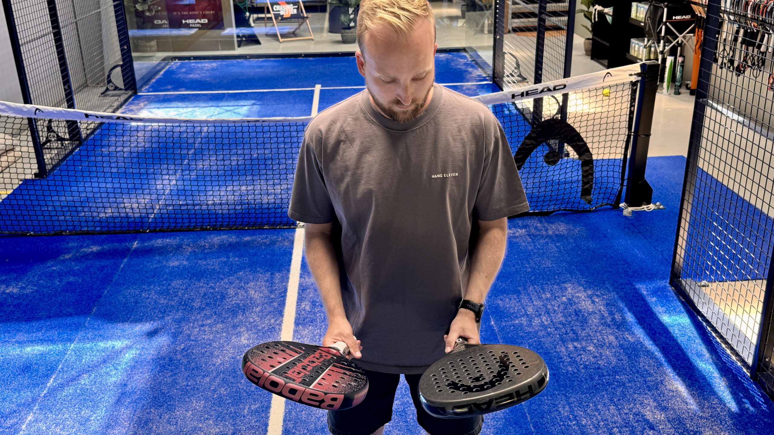 Mand holder en pink-sort Babolat padelbat og en sort HEAD padelbat op på padelbanen – sammenligning af begynderbat